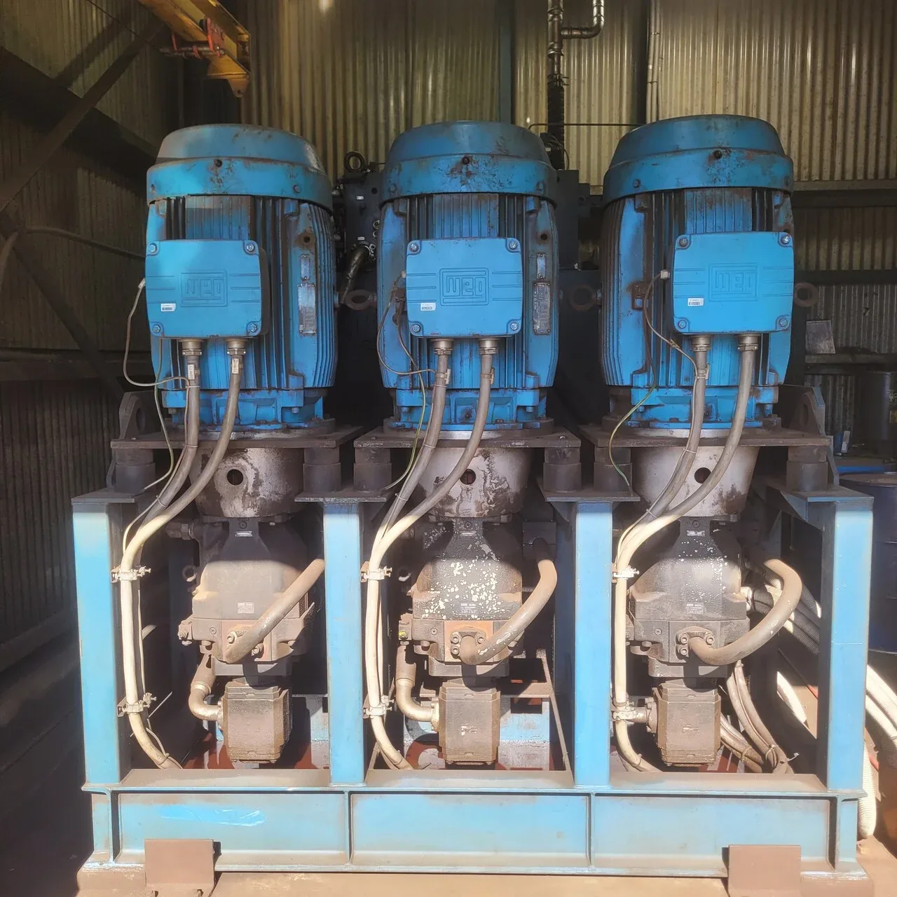 Three blue WEG electric motors mounted on vertical hydraulic pump units on a blue steel frame inside an industrial shed.