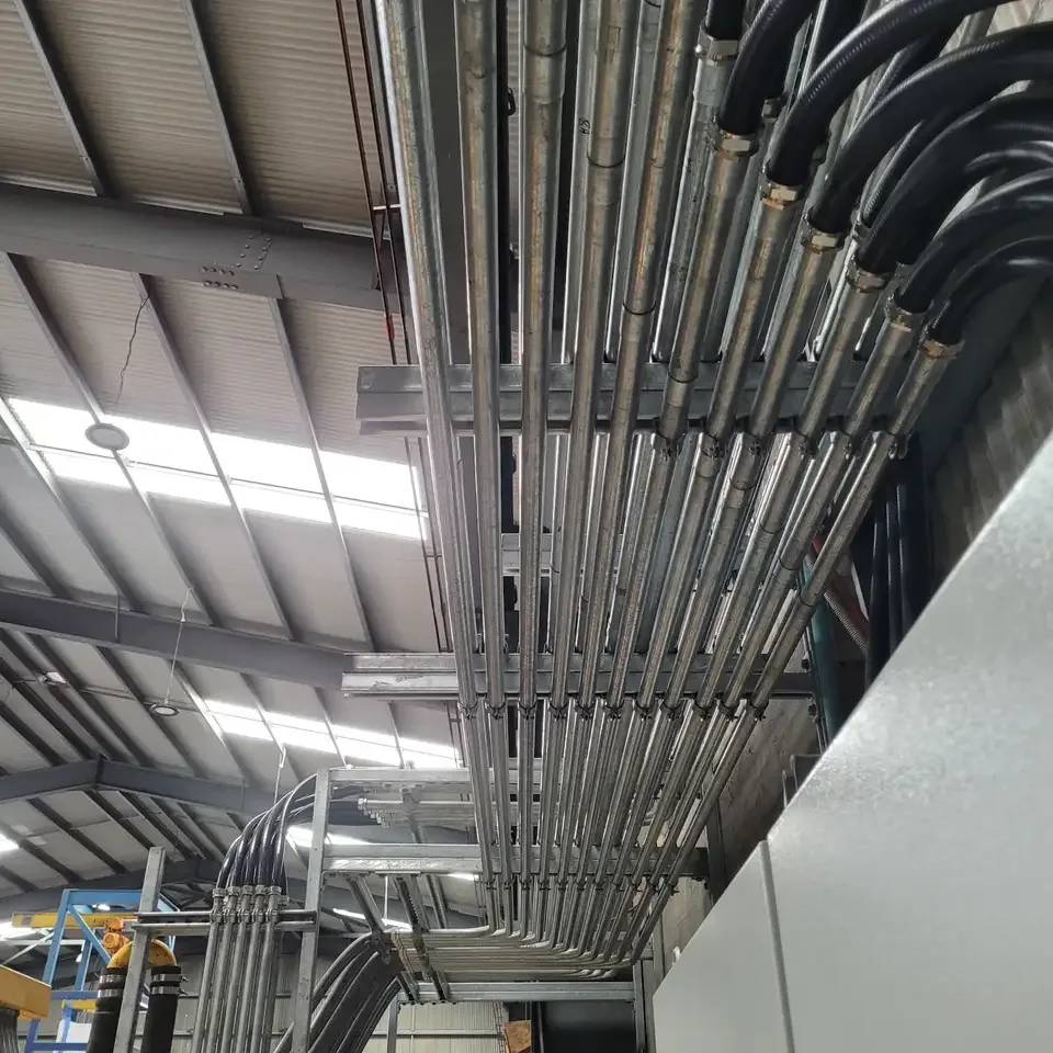 Galvanised steel conduit runs and black flexible conduit with brass glands fixed along a cable tray in an industrial building ceiling