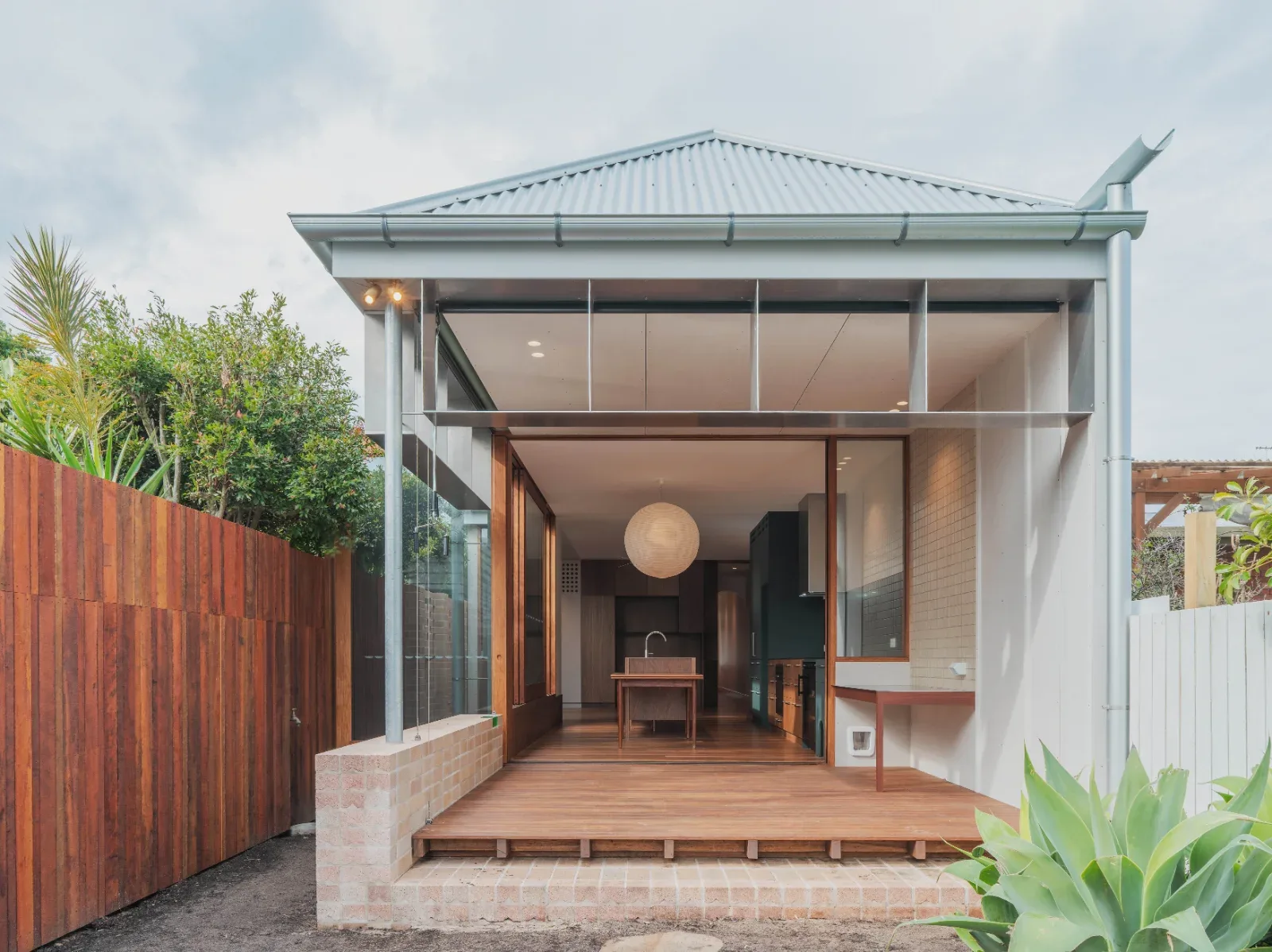 Renovated cottage with a corrugated metal gabled roof, a glass-walled extension and a timber deck, with the open kitchen visible through the sliding doors